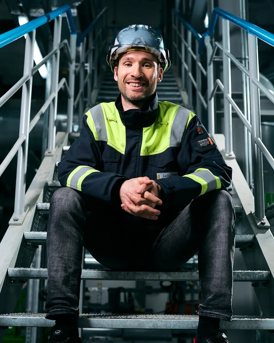 Mitarbeiter in Warnjacke und Helm sitzt lächelnd auf einer Industrietreppe