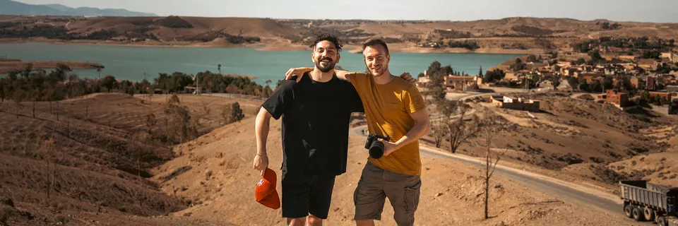 Zwei Crewmitglieder mit Equipment in einer Hügellandschaft am See